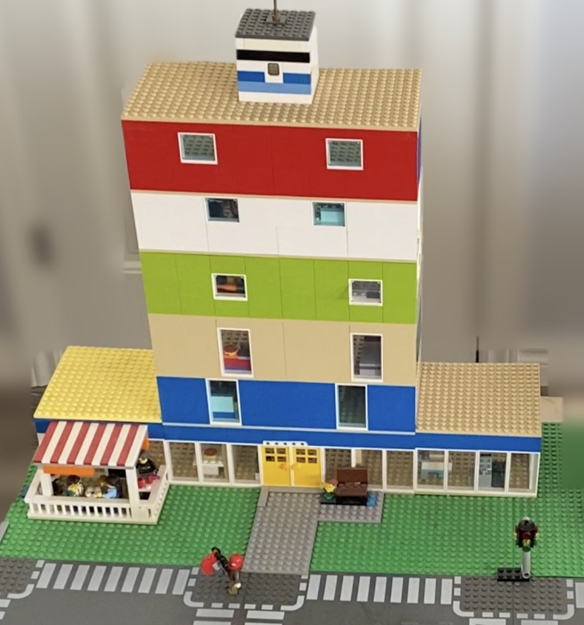 The original Mosaica: a Lego mixed-use building with shopfronts and a caf&eacute; patio at street level and four residential floors above, each in a different colour, set on a street corner with crosswalks.